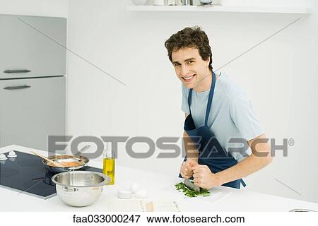 Stock Photo - Young man mincing basil in the kitchen, smiling at camera. Fotosearch