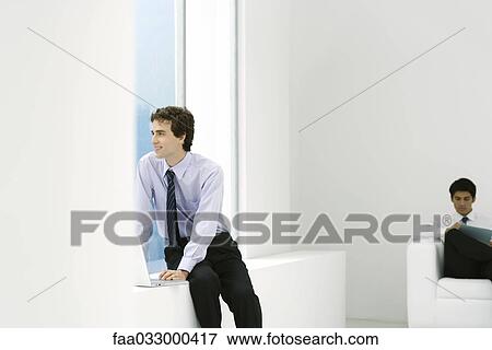 Stock Photo - Young professional sitting with laptop, looking out window, colleague reading in background. Fotosearch