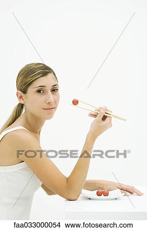 Picture - Young woman holding grape with chopsticks, looking over shoulder at camera. Fotosearch