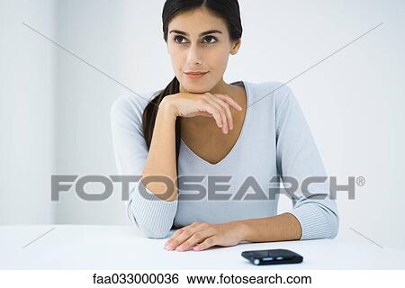 Stock Photograph - Young woman sitting with hand under chin, looking away, cell phone nearby. Fotosearch