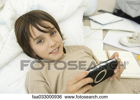 Stock Photograph - Boy holding handheld video game, lying down and smiling at camera. Fotosearch
