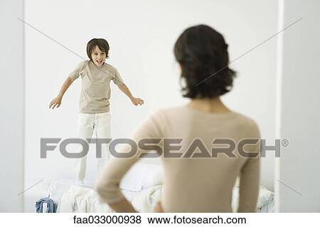 Stock Photo - Boy jumping on bed, mother standing in doorway with hand on her hip. Fotosearch