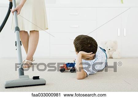 Stock Image - Boy lying on the ground with toys, looking up at his mother vacuuming around him. Fotosearch