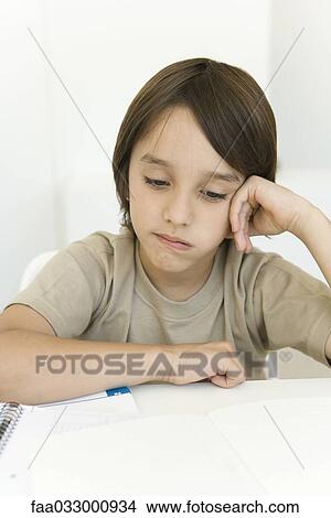 Boy sitting at table looking at homework, frowning View Large Photo Image Picture - Boy sitting at table looking at homework, frowning. Fotosearch