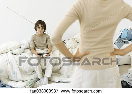 Stock Photo - Boy sitting in messy bedroom, holding handheld video game, mother in foreground with hands on hips, cropped. Fotosearch