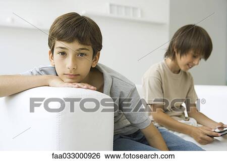 Boys sitting on couch, one looking at camera, the other playing handheld video game View Large Photo Image Stock Photograph - Boys sitting on couch, one looking at camera, the other playing handheld video game. Fotosearch