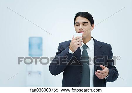 Stock Photography - Businessman holding and smelling disposable cup with eyes closed, water cooler in background. Fotosearch