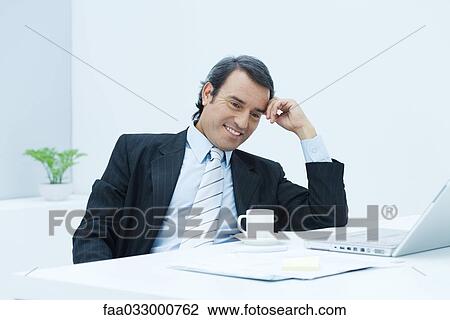 Stock Image - Businessman sitting at desk, leaning on elbow and holding head, looking down and smiling. Fotosearch
