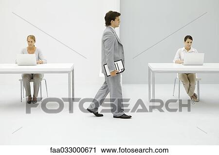Stock Image - Businessman walking in office, carrying document under arm, female colleagues working in background. Fotosearch