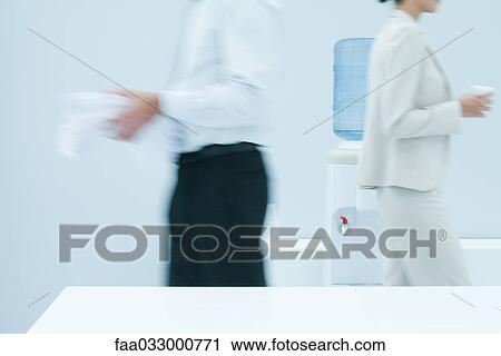 Stock Image - Colleagues walking in front of water cooler, blurred side view. Fotosearch