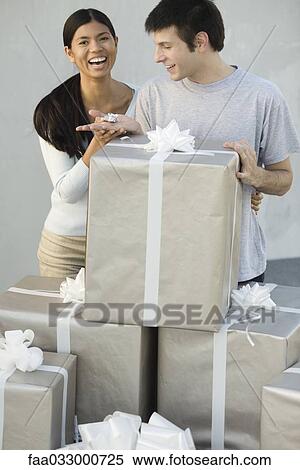 Stock Photography - Couple standing behind stack of large gift boxes, man holding tiny present, woman laughing. Fotosearch