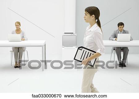 Stock Image - Female office worker carrying document under arm, colleagues working in background. Fotosearch