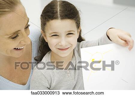 Stock Image - Girl holding up and showing drawing of the sun, mother smiling behind her daughter. Fotosearch