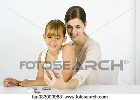 Stock Image - Girl sitting on young woman's lap, looking into hand mirror together. Fotosearch