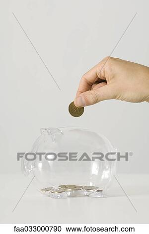 Stock Image - Hand dropping coin into transparent piggy bank. Fotosearch