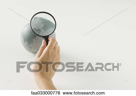 Stock Photo - Hand holding magnifying glass over globe, cropped view. Fotosearch