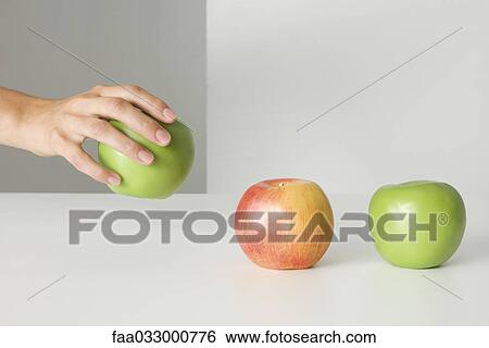 Stock Photograph - Hand picking up an apple, two other apples sitting in a row, cropped view. Fotosearch