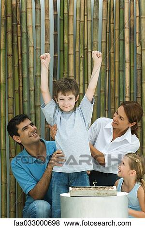 Stock Photo - Little boy standing beside potted seedling, arms raised, family smiling at him. Fotosearch