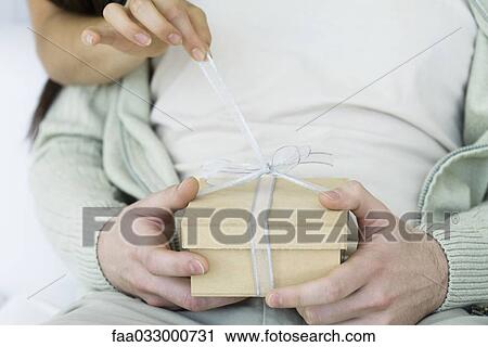 Stock Image - Man holding gift, woman's arm reaching around to untie the ribbon, cropped view. Fotosearch