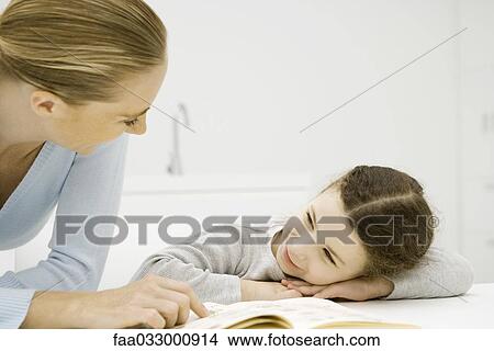 Mother and daughter smiling at each other, girl resting head on arms, woman pointing at book View Large Photo Image Picture - Mother and daughter smiling at each other, girl resting head on arms, woman pointing at book. Fotosearch