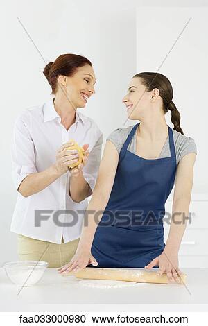 Stock Image - Mother and teen daughter baking together, smiling at each other. Fotosearch
