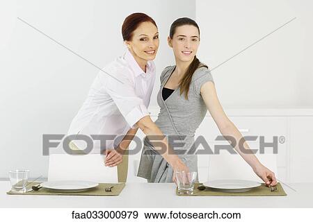 Stock Photo - Mother and teen daughter setting the table together, smiling at camera. Fotosearch
