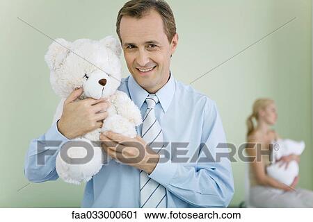 Stock Image - New father holding teddy bear, smiling at camera, wife holding baby in background. Fotosearch