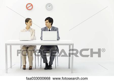 Stock Photo - Office workers sitting side by side beneath clocks, looking at each other. Fotosearch