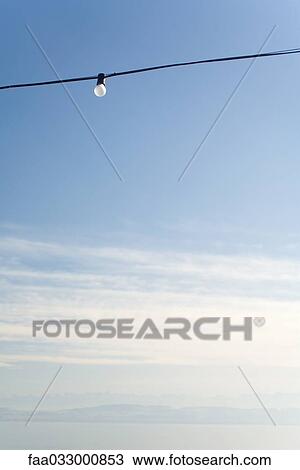 Stock Image - Power line with a single light bulb, cloudy sky, low angle view. Fotosearch
