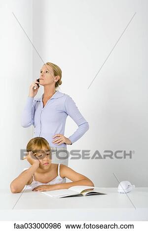 Preteen girl sitting at table with homework, mother standing behind her, using cell phone View Large Photo Image Stock Photograph - Preteen girl sitting at table with homework, mother standing behind her, using cell phone. Fotosearch