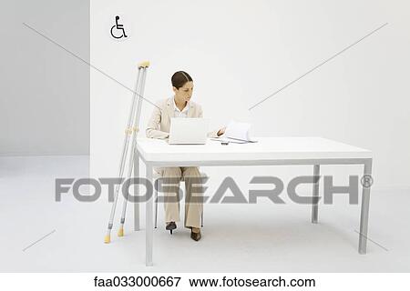 Stock Photo - Professional woman working in office, crutches leaning against desk beside her. Fotosearch