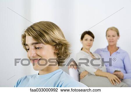 Stock Image - Teenage boy looking away, women listening to MP3 player in background, cropped view. Fotosearch