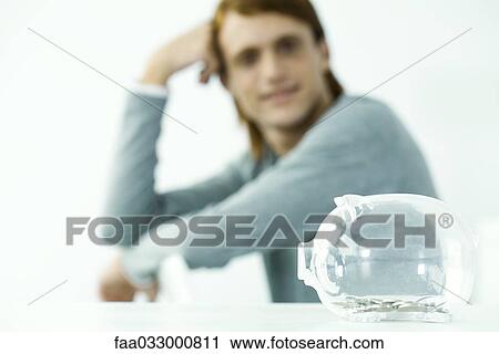 Stock Image - Transparent piggy bank sitting on table, young man in background holding head. Fotosearch