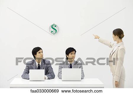 Stock Photo - Two male office workers sitting at desk, female supervisor pointing at clock with dollar sign. Fotosearch