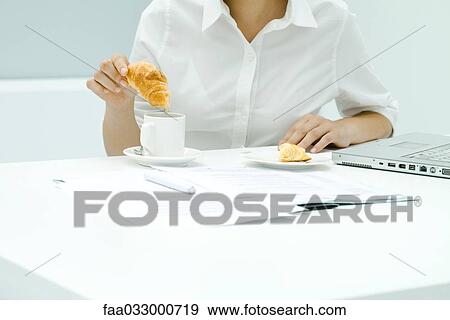 Stock Photo - Woman dipping croissant into coffee, sitting at cluttered desk, cropped view. Fotosearch