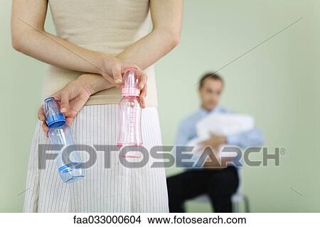 Woman holding baby bottles behind her back, husband holding baby in background, cropped view View Large Photo Image Picture - Woman holding baby bottles behind her back, husband holding baby in background, cropped view. Fotosearch