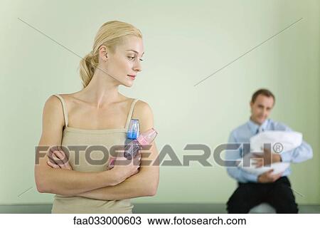 Stock Image - Woman holding baby bottles, looking over shoulder at husband holding baby in background. Fotosearch