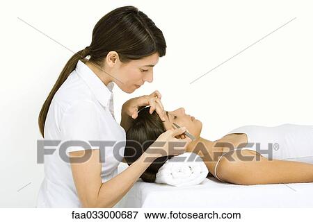 Stock Photo - Woman lying on back, having eyebrows tweezed, side view. Fotosearch
