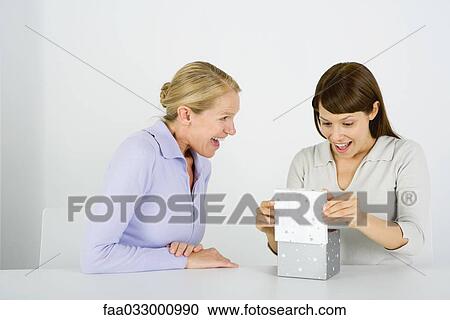 Stock Image - Woman opening gift, another woman sitting nearby watching, both excited. Fotosearch