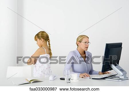 Woman sitting at table using desktop, daughter standing nearby with her back turned and arms folded View Large Photo Image Stock Photo - Woman sitting at table using desktop, daughter standing nearby with her back turned and arms folded. Fotosearch