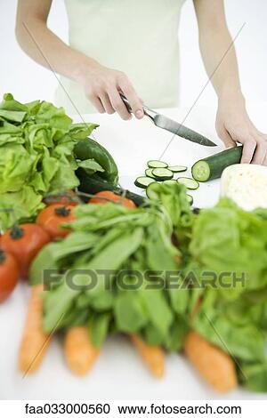 Stock Image - Woman slicing cucumber, assorted vegetables in foreground, cropped view. Fotosearch