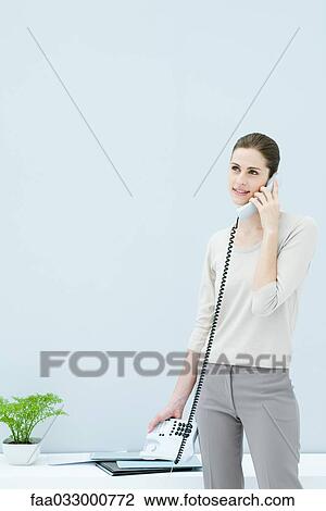 Stock Image - Woman standing and talking on landline phone, looking away and smiling. Fotosearch