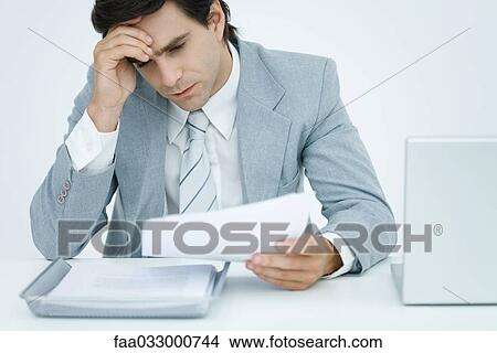 Picture - Young businessman sitting at desk, holding head and looking down at document. Fotosearch