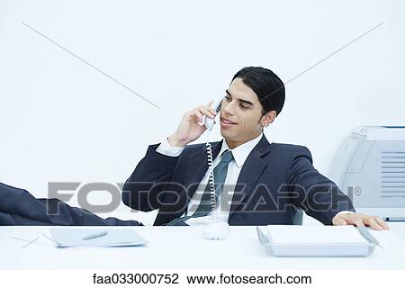 Stock Image - Young businessman sitting with feet up at desk, talking on landline phone. Fotosearch