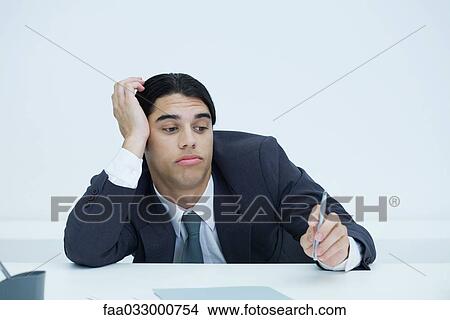 Picture - Young businessman slouching at desk, holding head and looking at pen with bored expression. Fotosearch