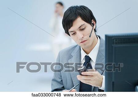 Stock Photo - Young businessman wearing headset and looking down at cell phone. Fotosearch