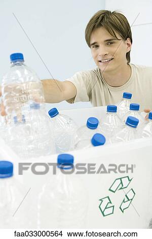 Picture - Young man placing plastic bottle in recycling bin, smiling at camera. Fotosearch