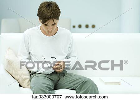 Stock Photo - Young man sitting on couch, cleaning glasses with shirt. Fotosearch