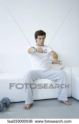 Stock Photograph - Young man sitting on sofa, lifting dumbbells. Fotosearch
