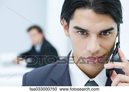Stock Image - Young professional man holding cell phone, smirking at camera, portrait. Fotosearch